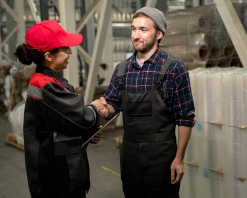 tuotekorttikuva jossa kaksi ihmistä kättelevät teollisuushallissa työasut yllään.