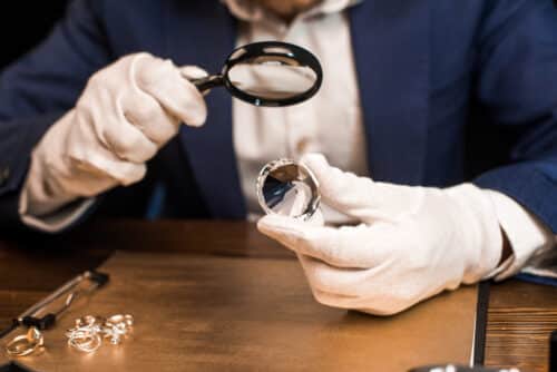 Henkilö arvioi timanttia suurennuslasin avulla valkoiset käsineet käsissään. Tuotekuva.